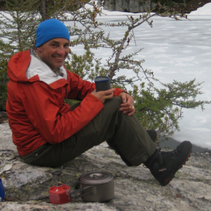Mark Peyron sitting on a rock, snow in background, wearing blue hat, red jacket, dark olive pants and hiking boots
